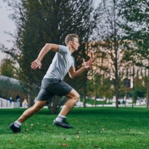 A man, dressed in a gray t-shirt and dark shorts, runs effortlessly on the grass in a park. Black shoes traction beneath him as trees and buildings form the backdrop, evoking the vitality one might seek from a nearby sports medicine doctor in Miller Place.