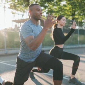 On a sunny day in East Setauket, NY, two people perform lunges outdoors, embodying the principles of sports medicine with trees and a fence offering a serene backdrop.