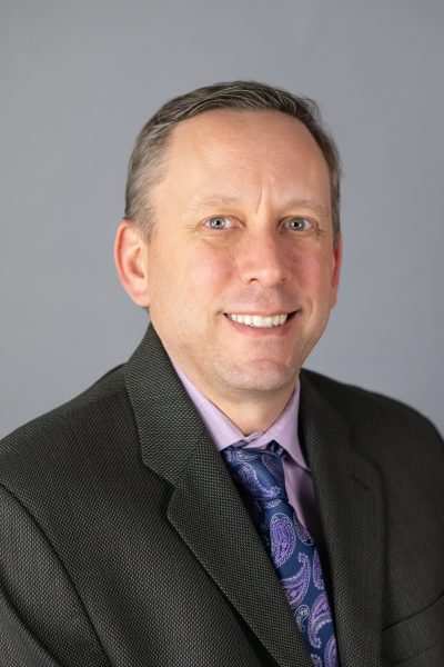 A man in a dark blazer and patterned tie poses against a gray background, smiling slightly.