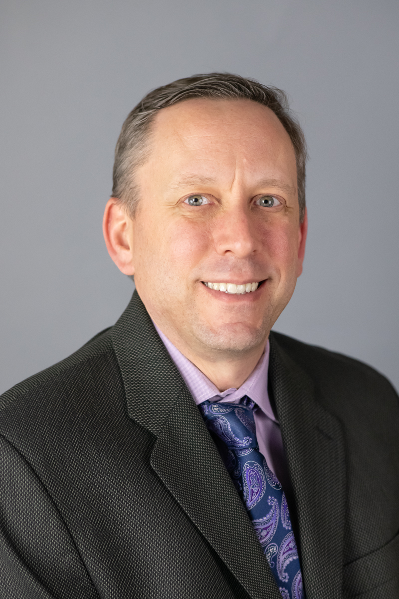A man in a dark blazer and patterned tie poses against a gray background, smiling slightly. A man in a dark blazer and patterned tie poses against a gray background, smiling slightly